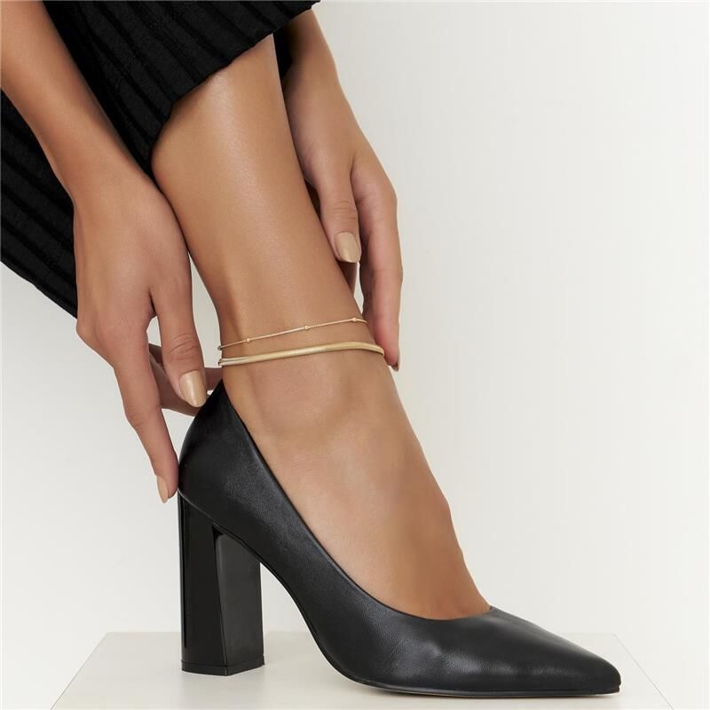Modelo usando tornozeleira Lizza dourada, semijoia antialérgica de acabamento liso e tecnologia antialérgica, destacada com sapato preto elegante.