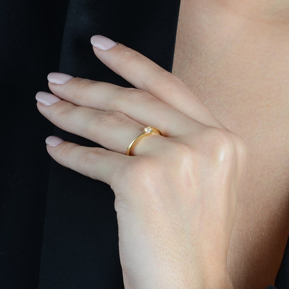 Anel dourado solitário com zircônia, semijoias de alta qualidade em 18k e 24k antialérgicas, em mão elegante sobre tecido preto.